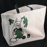 The Fox Project Tote by Mum n Sun Ink from Bonnie Bishoff is a beige Envirotote cotton/poly canvas bag with black handles, showcasing an illustration of a fox knitting with green yarn. The fox is encircled by green pine branches against a black background. The tote also features two side pockets and is sewn in New Hampshire.