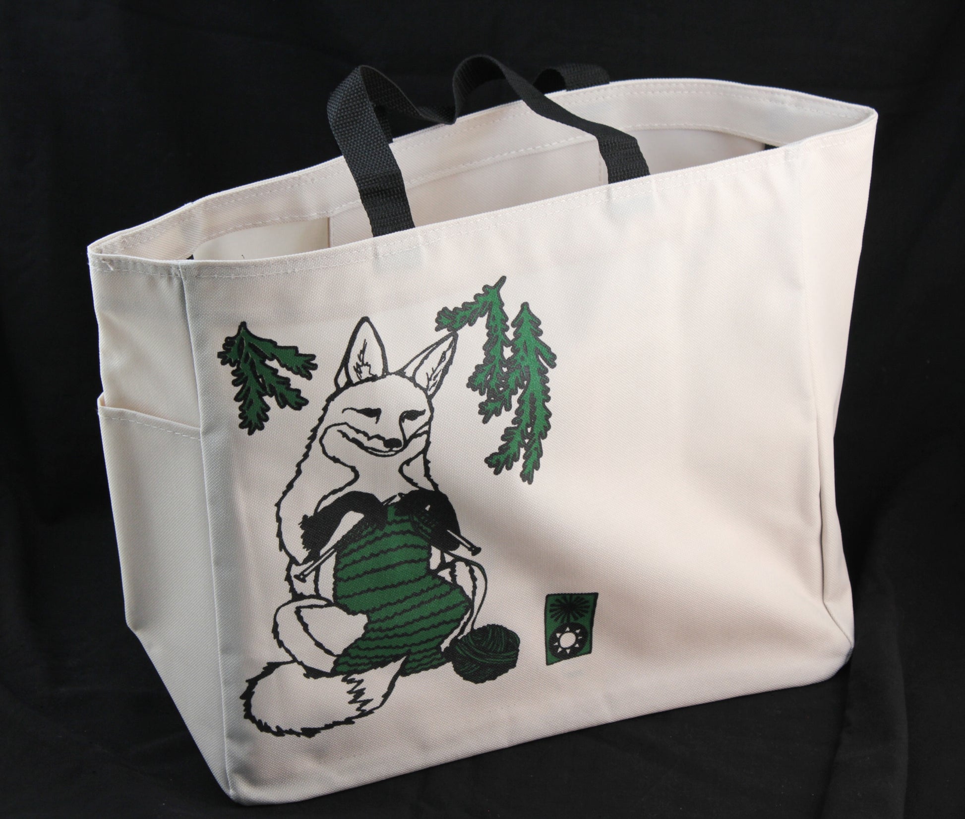 The Fox Project Tote by Mum n Sun Ink from Bonnie Bishoff is a beige Envirotote cotton/poly canvas bag with black handles, showcasing an illustration of a fox knitting with green yarn. The fox is encircled by green pine branches against a black background. The tote also features two side pockets and is sewn in New Hampshire.