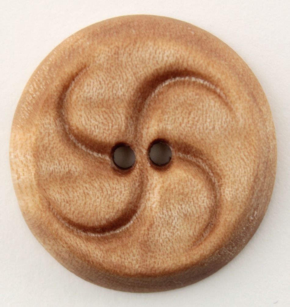 A close-up image of the Wood Button by Alosada, crafted by Alosada LLC, shows a round, light brown wooden button made from local Maine wood. The button features two small holes in the center and a carved, wavy swirl pattern that gives it a textured appearance. The visible wood grain adds to its rustic charm, emphasizing its all-natural local materials.