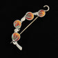 The Spring Bud Shawl Pin by Bonnie Bishoff Designs, crafted by Bonnie Bishoff, boasts a silver metal base with a secure locking pin on one end and features four round enamel discs. Each disc showcases a vibrant floral design in hues of orange, red, green, and blue. Ideal for adding charm to knitted wearables, this brooch is beautifully set against a black background.