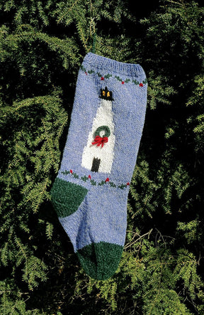 The Maine Christmas Stocking Kits by Bully Woolies present a stocking that evokes the charm of a family heirloom, featuring a lighthouse with a wreath on a light blue background. Made from locally-spun Maine wool yarn, its toe and heel are dark green and embellished with cheerful red and green details. It drapes elegantly against a vibrant, green bush.