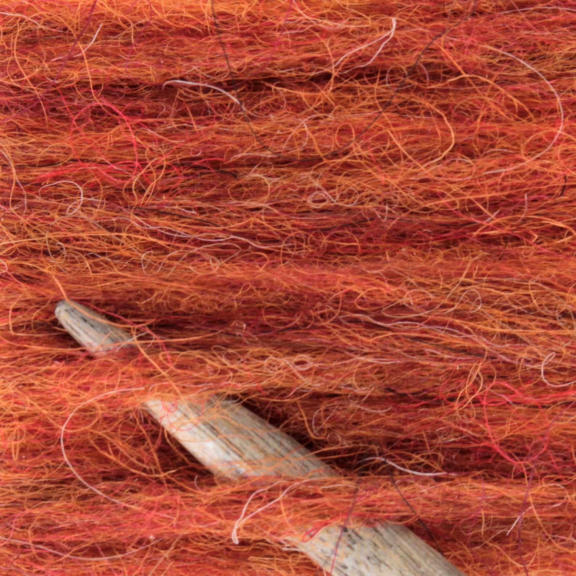 Close-up of vibrant orange and red Berroco Plötulopi Unspun Icelandic Wool Yarn with specks of different shades. A wooden knitting needle is partially visible nestled within the unspun yarn, showcasing the texture and fibers of these Plötulopi materials.