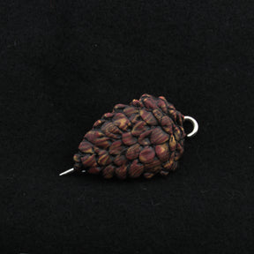 A close-up photograph shows the Woodland Shawl Pin by Bonnie Bishoff Designs, a brown pine cone ornament against a black background. This lightweight accessory by Bonnie Bishoff features a shiny, metallic hook at its top, indicating that it can be hung as decoration or used in polymer clay designs.