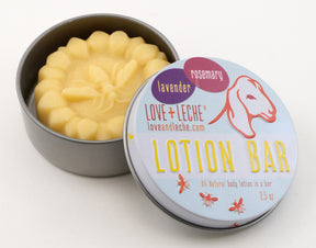 An open tin containing a round lotion bar with a sunflower design on top. The tin lid beside it reads "LOVE + LECHE, Lavender Rosemary, Love + Leche Lotion Bar, All Natural body lotion in a bar with beeswax, 2.5 oz." The lid also features a goat illustration and a website URL.