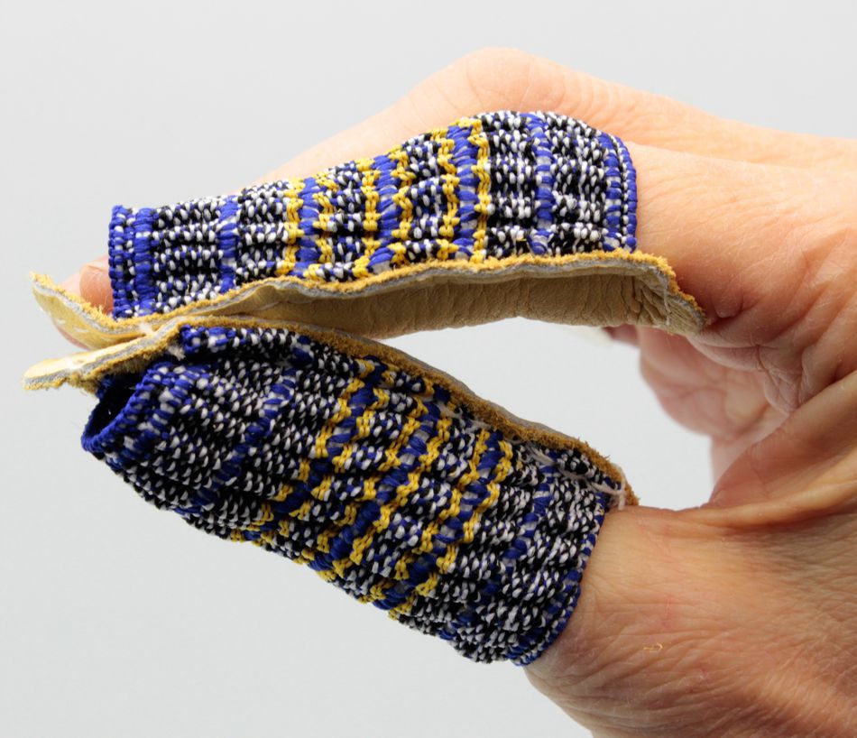 Two fingers are shown wearing *Leather Finger Guards, Open-End* from Magid Glove & Safety Co LLC, featuring colorful, hand-woven patterns in shades of blue, yellow, and white. These fabric protectors come with a leather backing and are perfect for sewing or quilting enthusiasts. The fingers are held close together against a plain grey background.