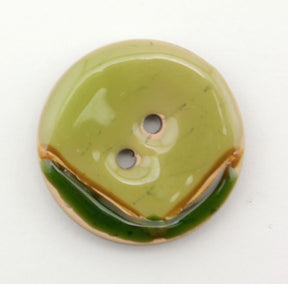 The Coco Boomerang 1 1/8" Button by Buttons Etc. is a round, glossy green button with two holes in the center. The upper half is a lighter shade of green, while the lower half features a darker green curved line resembling a smile. Reminiscent of colorful shellac designs, this button also includes small marbled brown sections within it.