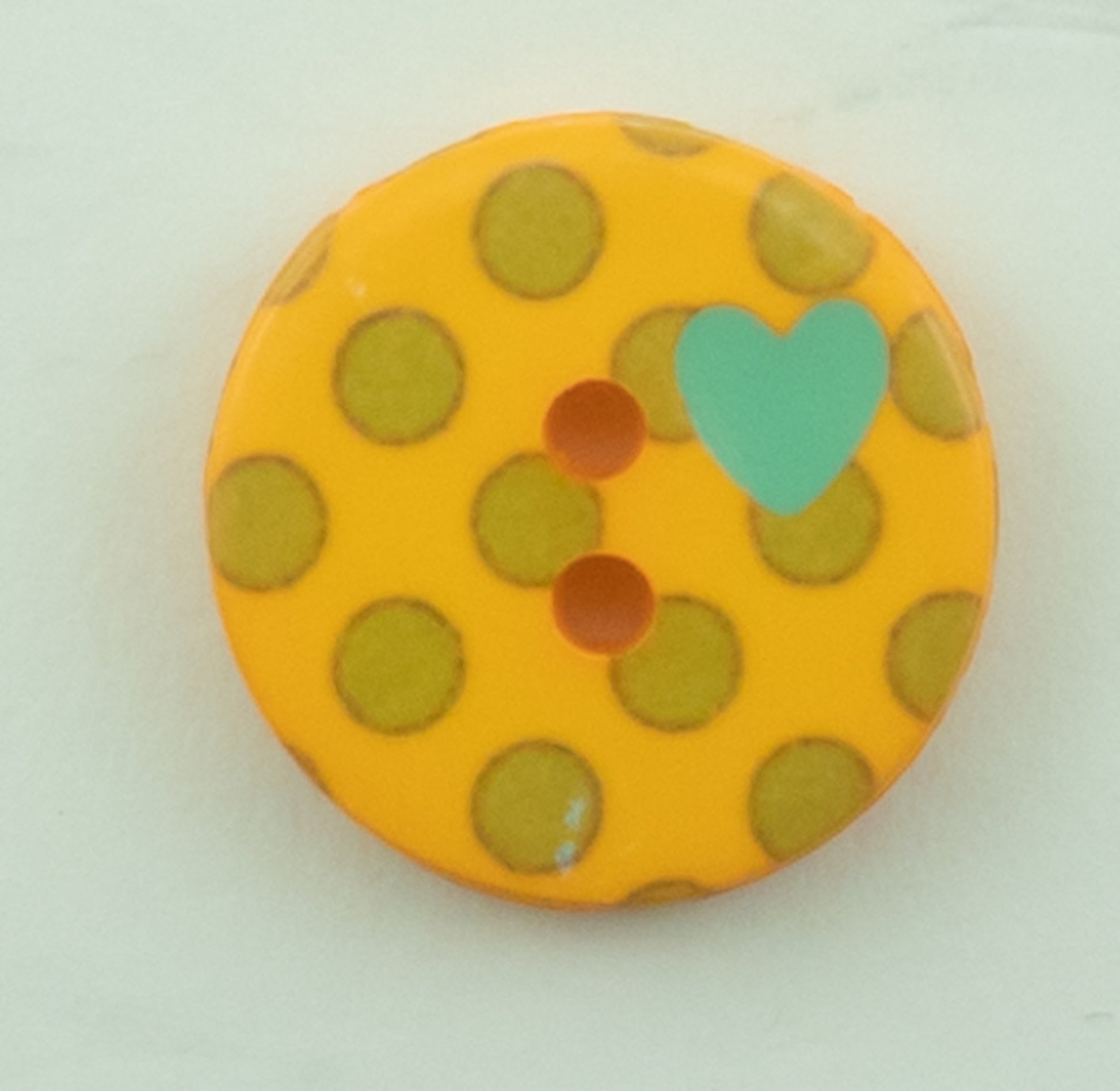 A round 5/8" diameter Dots Button from Buttons Etc., made of orange plastic, with green polka dots and two holes in the center. It features a small teal heart shape printed near the edge and is displayed against a plain, light background.