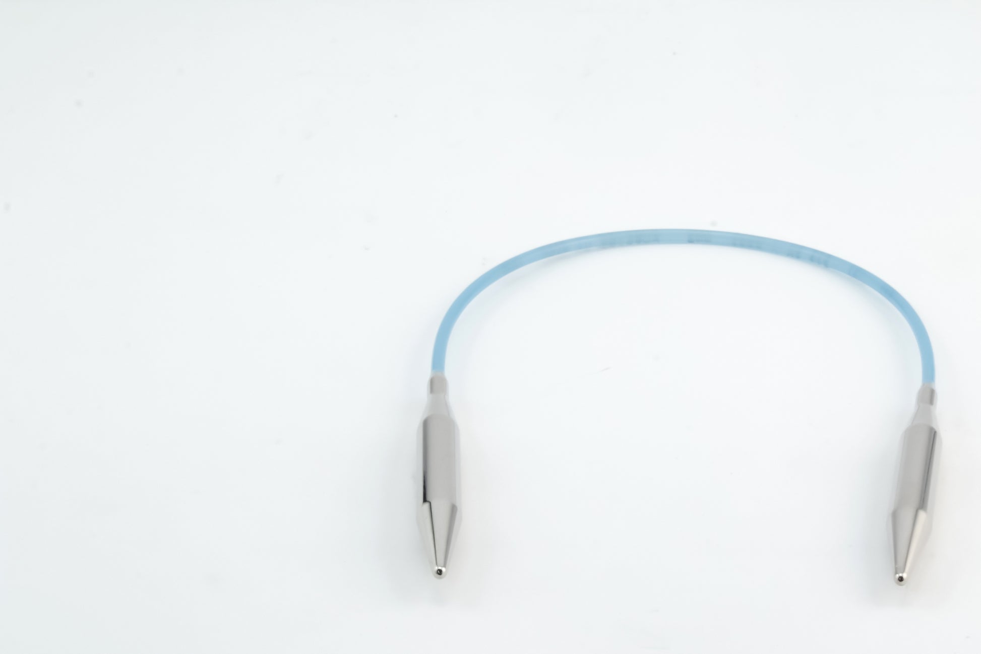 A flexible blue cable with metallic connectors on both ends is laid out against a white background. The sleek, cylindrical connectors suggest it could be Addi Turbo Circular Knitting Needles by Skacel, possibly designed for electronic devices or specialized uses like knitting cuffs and collars on doll clothes.