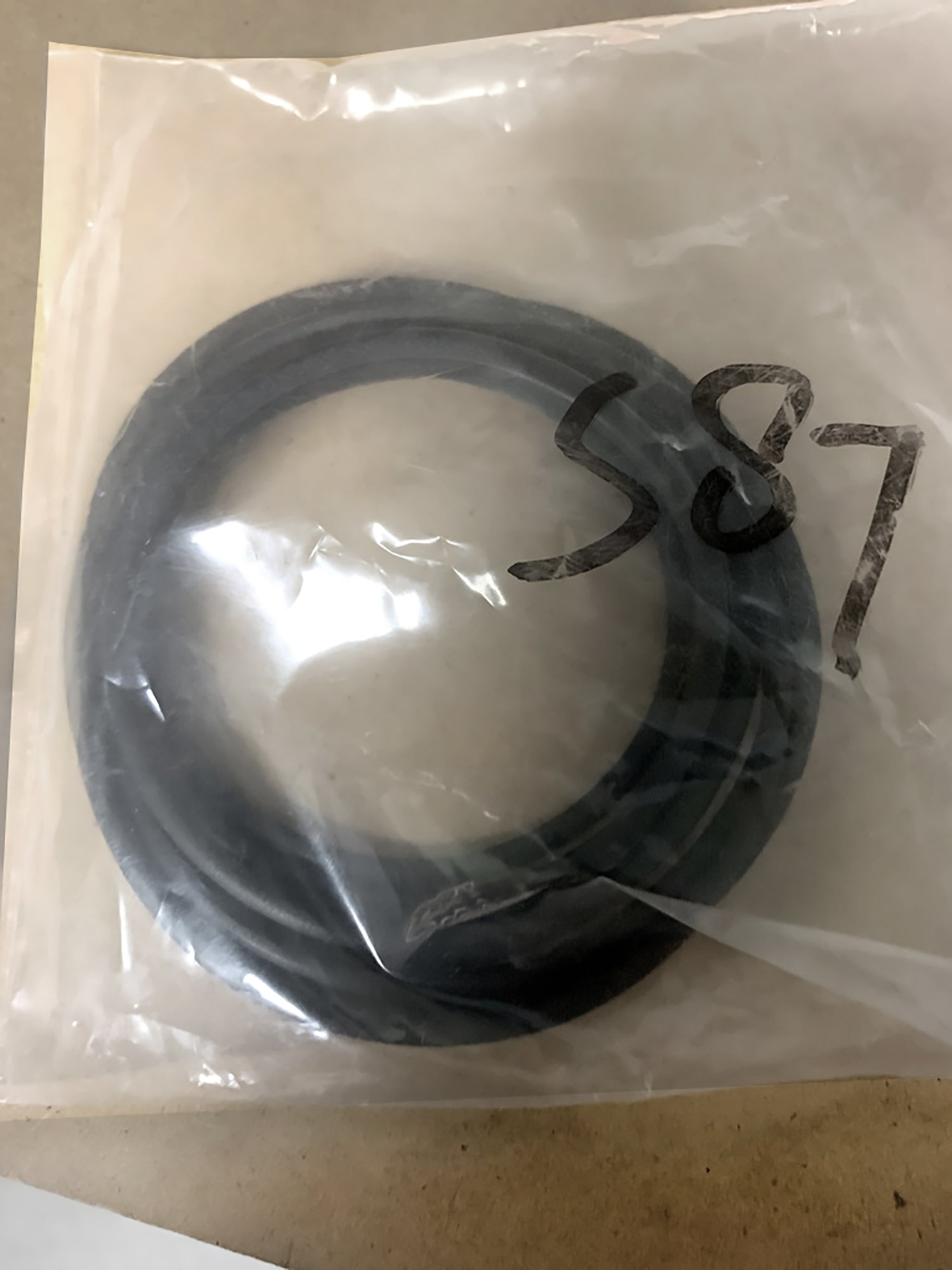 A sealed plastic bag contains a black rubber ring, which is a drive band for a Louët spinning wheel. The bag is labeled with handwritten text reading "S87" and sits on a tan surface. The product is part of the Louët Spinning Wheel Drive Bands/Belts line from Louët Inc.