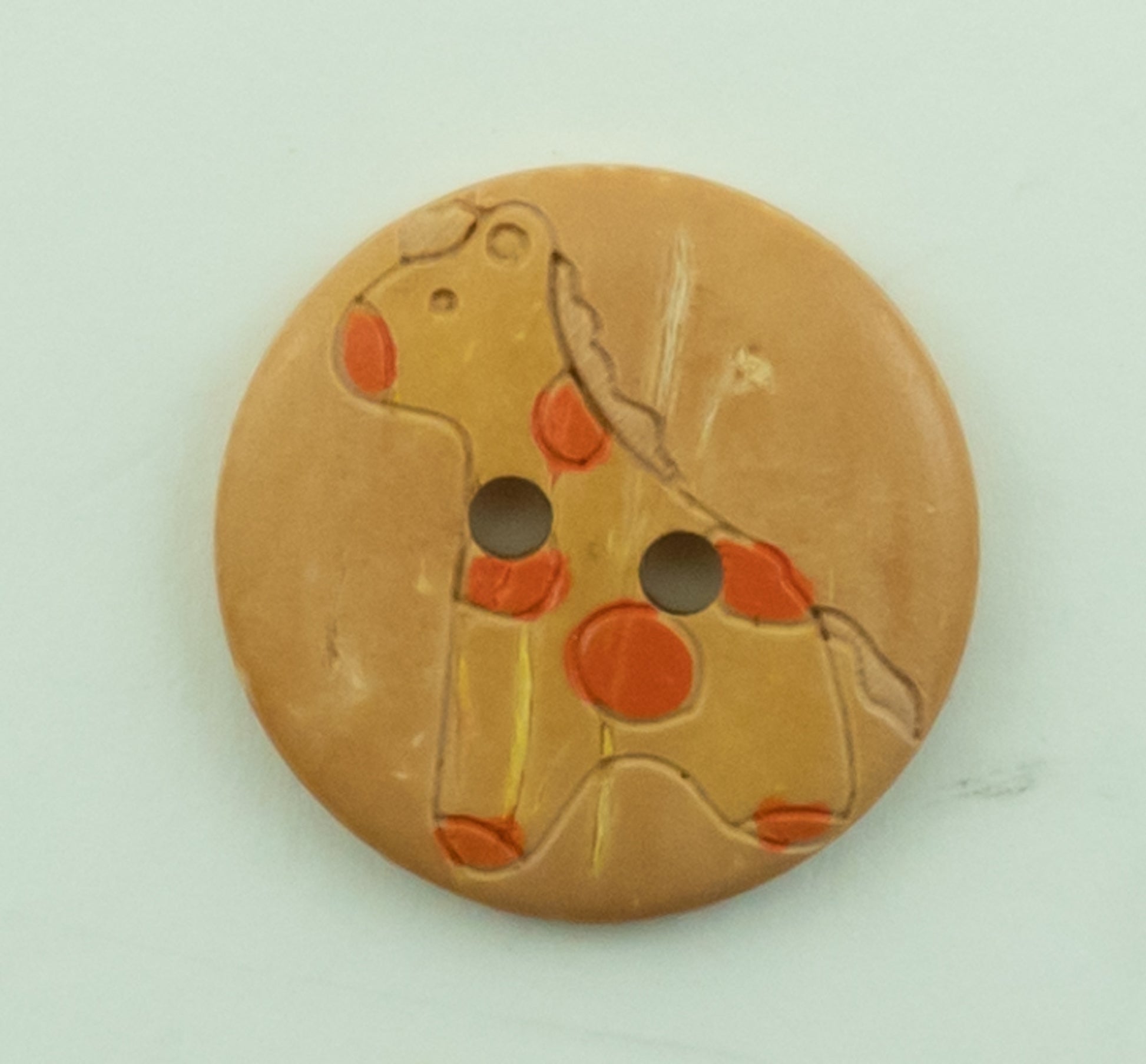 The Baby Button by Buttons Etc. is a 13/16" diameter, round, light brown button featuring an engraved and painted design of a giraffe. The giraffe is outlined with red and orange spots and has two holes in the middle. The button's surface shows signs of slight wear.