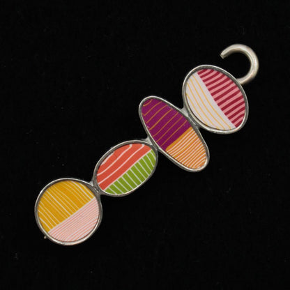 The Bubbles Shawl Pin by Bonnie Bishoff Designs features four polymer clay ovals set against a black background. Each oval displays a unique color and pattern: red and white stripes, purple and gold stripes, green and orange stripes, and yellow and pink stripes. A hook is attached at the top for secure placement.