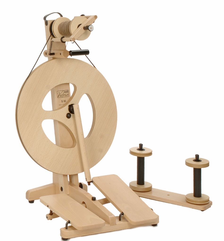 The Louët Victoria Double-Treadle Folding Spinning Wheel is a wooden spinning wheel with a simple, modern design, perfect for spinning yarn. It features a large wheel with three oval cutouts, dual treadles, and an adjustable bobbin brake setup for holding yarn. Additional spools and a high-speed set are displayed beside the main unit. The brand name "Louët Inc." is visible on the wheel.