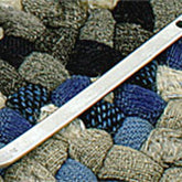 A silver steel tent stake with two holes near the top lies on a textured, woven surface composed of various shades of blue, gray, and brown fabric, reminiscent of patterns created with Whitman Tool & Die Co., Inc.'s Braidkin Lacer.