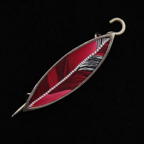 The Songbird Shawl Pin by Bonnie Bishoff Designs, crafted by Bonnie Bishoff, is a handmade metal brooch shaped like a leaf with a striking red and white striped pattern set against a black background. It features a secure locking pin for easy attachment, making it the perfect gift.