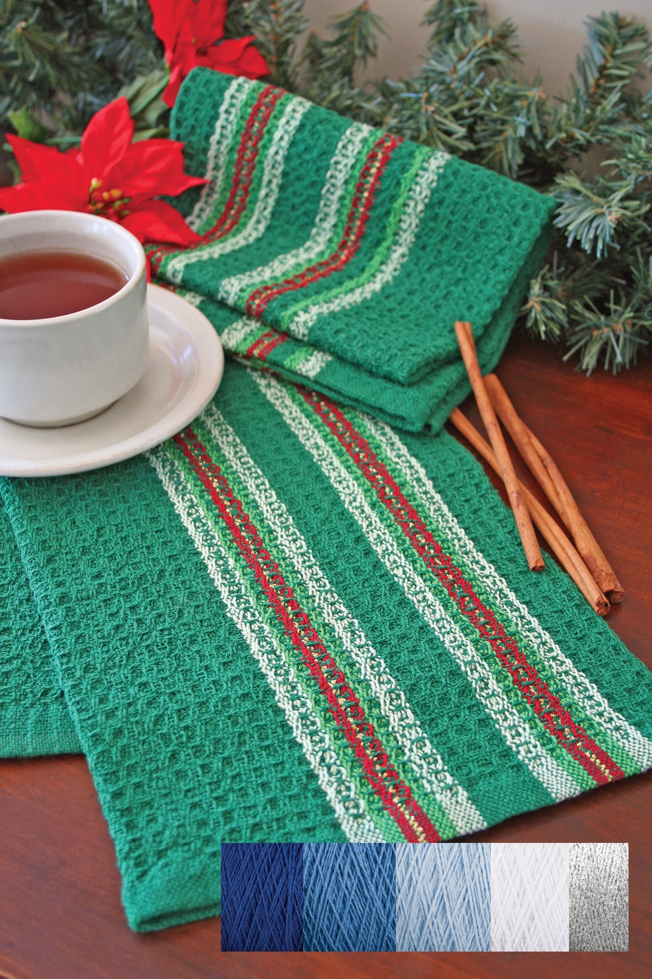 Halcyon Yarn's Waffle Weave Dish Towel Kit features green woven dish towels with red, white, and green stripes displayed on a wooden surface. A white cup filled with tea sits on one towel, surrounded by cinnamon sticks and red poinsettias. Below, there is a palette of yarn options in blue, green, grey, and white for your weaving kit.