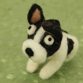 A small, handmade figurine of a black and white dog made from felt, featuring wide, expressive eyes and perky ears with one ear showing a patch of brown. The figurine is crafted using the Woolbuddy Needle Felting Kits by Woolbuddy and is placed on a green textured background.