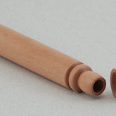 A cylindrical Beech Wood Needle Case from Bryson Distributing, Inc. with a removable cap lies on a white surface. The case appears hollow and has smooth, light-colored wood with a simple design. This wooden tapestry needle case features a cap placed next to it, revealing its rounded interior.