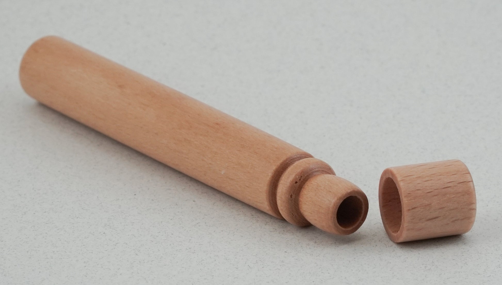 A cylindrical Beech Wood Needle Case from Bryson Distributing, Inc. with a removable cap lies on a white surface. The case appears hollow and has smooth, light-colored wood with a simple design. This wooden tapestry needle case features a cap placed next to it, revealing its rounded interior.