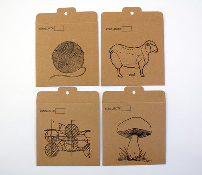 The Halcyon Yarn Interchangeable Needle Cord Envelopes come in a set of four kraft paper envelopes, each labeled "CORD LENGTH." They feature black line drawings of a ball of yarn, a sheep with dotted lines on its body, geometric shapes, and a mushroom. Each envelope has a tab at the top with a small hole for hanging, making them perfect for organizing travel notions or interchangeable cords.