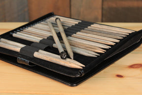 A Lykke Single-Point Knitting Needle Set, 10" by Knitting Fever / Euro Yarns is open on a wooden surface, displaying an array of knitting needles neatly organized in individual elastic loops. Two knitting needles are prominently placed on top of the others. The background features a wooden panel.