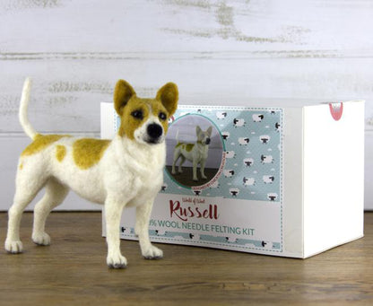 A needle felted dog sculpture stands on a wooden surface in front of a box. The box, labeled "Russell," features an image of the realistic animal and includes information about a "100% wool Animal Sculpture Felting Kit" by Europa Wools Ltd. The background is decorated with small sheep patterns.