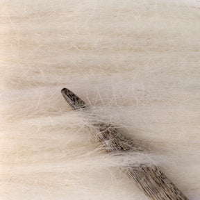 A close-up image of a wooden knitting needle resting on soft, off-white Berroco Plötulopi Unspun Icelandic Wool Yarn. The image showcases the texture of both the needle and the yarn, highlighting the intricate details and natural fibers.