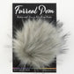 A package labeled "Furreal Pom by KFI" from Knitting Fever / Euro Yarns displays a fluffy, gray faux fur pompom attached to a black backing card. The text on the packaging reads "Extra-soft Vegan Fur Pom Poms" and "KFI Collection." The pompom has a soft, wispy texture with varying shades of gray.