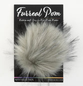 A package labeled "Furreal Pom by KFI" from Knitting Fever / Euro Yarns displays a fluffy, gray faux fur pompom attached to a black backing card. The text on the packaging reads "Extra-soft Vegan Fur Pom Poms" and "KFI Collection." The pompom has a soft, wispy texture with varying shades of gray.