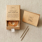 A Cocoknits Leather Cord and Needle Stitch Holder Kit is presented. The kit, which includes stitch holders with a leather cord and two knitting needles, is placed in a small, open cardboard box. A smaller box with the message "delight in the making" is also included. The packaging is minimalist and eco-friendly.