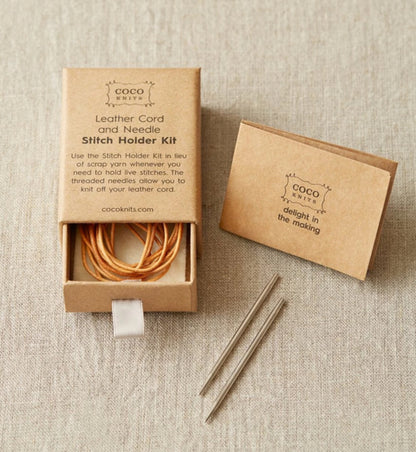 A Cocoknits Leather Cord and Needle Stitch Holder Kit is presented. The kit, which includes stitch holders with a leather cord and two knitting needles, is placed in a small, open cardboard box. A smaller box with the message "delight in the making" is also included. The packaging is minimalist and eco-friendly.