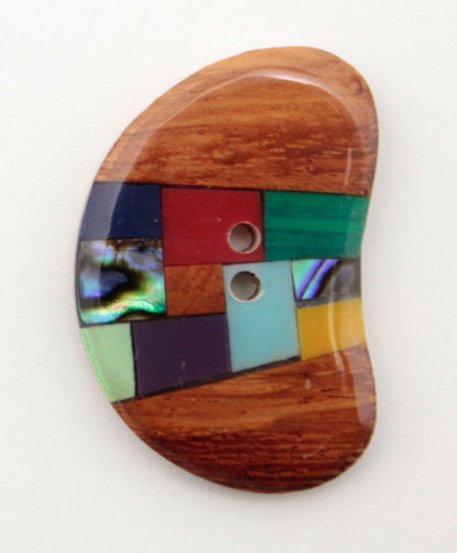 The Wood Gems Shell 1 3/8" Button by Buttons Etc. is a uniquely shaped wooden button with a curved, bean-like form and two holes. This oblong button features a vibrant inset of colorful geometric tiles, including shades of turquoise, purple, green, blue, and red, arranged in a mosaic pattern.