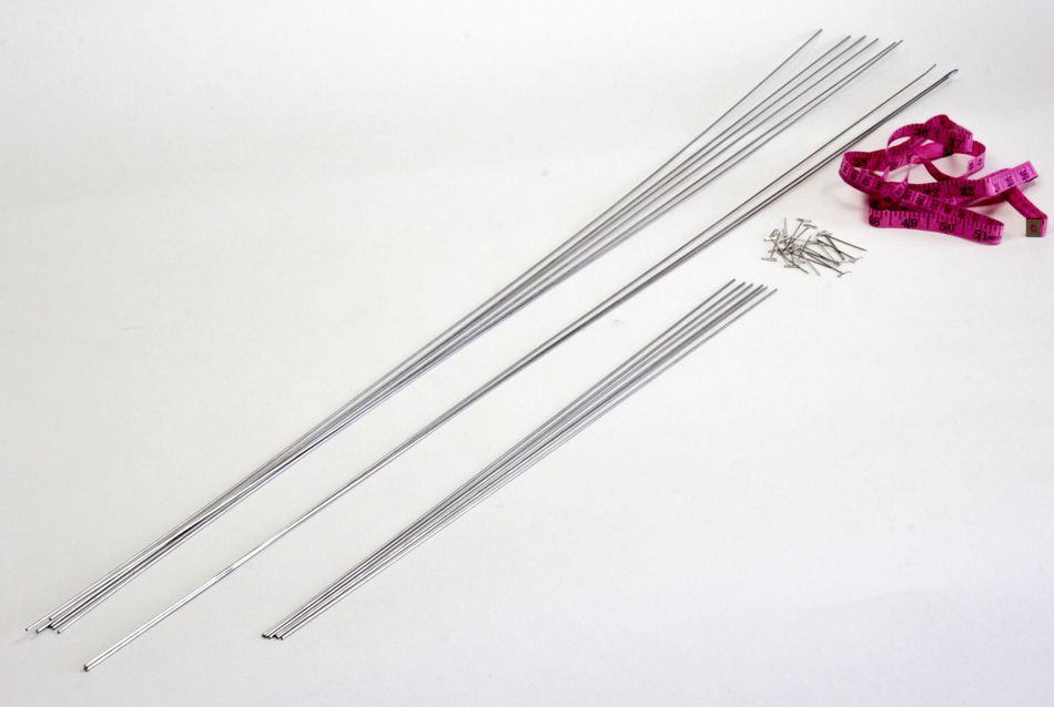 Lace Blocking Wire Kit by Knitter's Pride from Accessories Unlimited arranged on a white surface, featuring several long stainless steel knitting needles of various sizes, a small collection of short metal pins, and a pink measuring tape neatly coiled to the side.