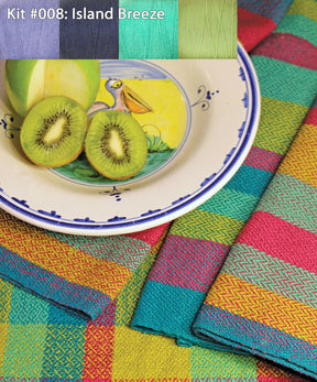 A vibrant Organic Cottolin Tea Towel Kit from Halcyon Yarn, labeled "Kit #008: Island Breeze," displays colorful threads and fruits. The fabric, suitable for a 4 shaft loom, features bright patterns in teal, yellow, pink, and green. A plate with a pelican illustration holds a sliced kiwi and a partially cut green apple.