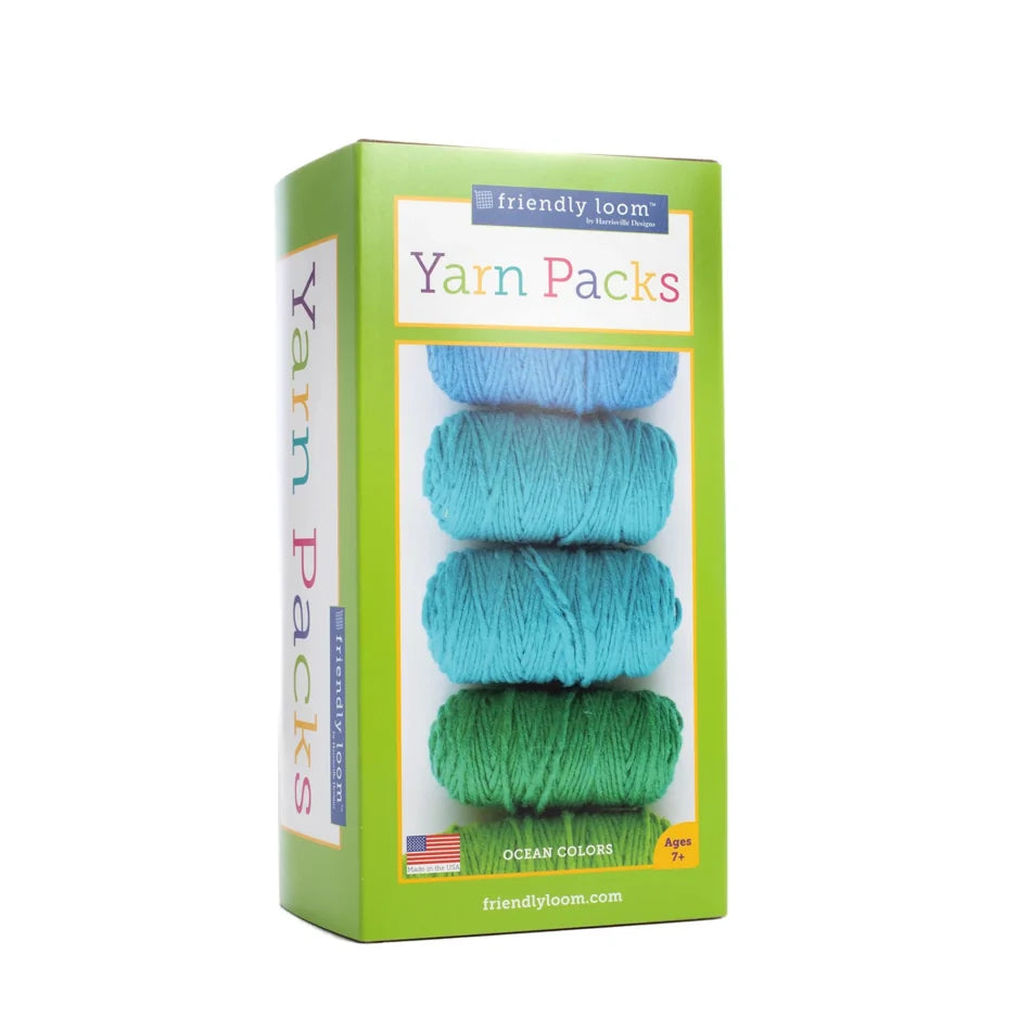 The image shows a package of "Friendly Loom Lap Loom Yarn Pack" containing six balls of 100% wool yarn in shades of blue and green, labeled as "Ocean Colors." Suitable for ages 7 and up, it is made in the USA. Ideal for use with a peg loom, the packaging is green with colorful text.